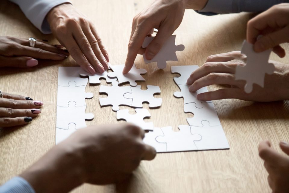 Hands of seven people sitting at wooden table putting together a puzzle