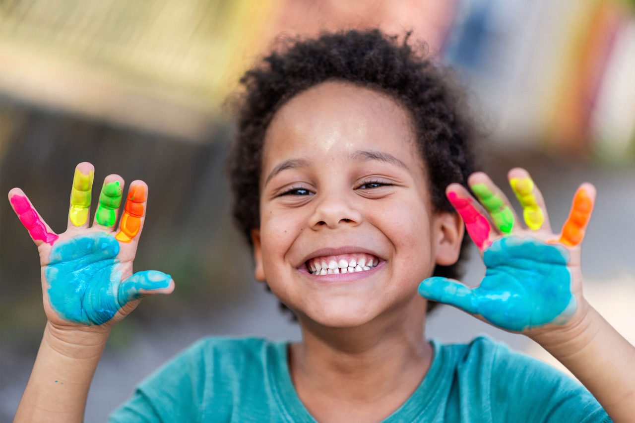 A smiling young boy holds up his hands which are covered in paint