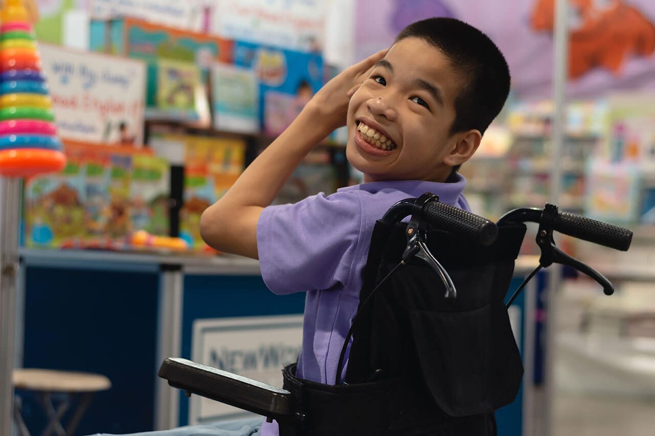 A young boy in a wheelchair smiles in front of a rack of children's books