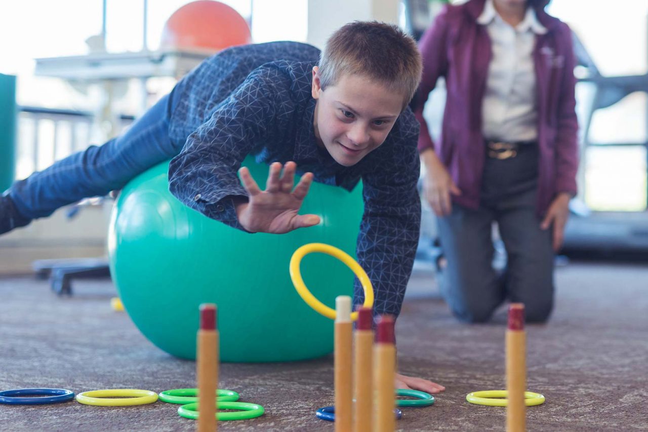 adolescent boy in blue button up shirt laying across large teal exercise ball tossing plastic yellow ring onto ring toss for occupational therapy