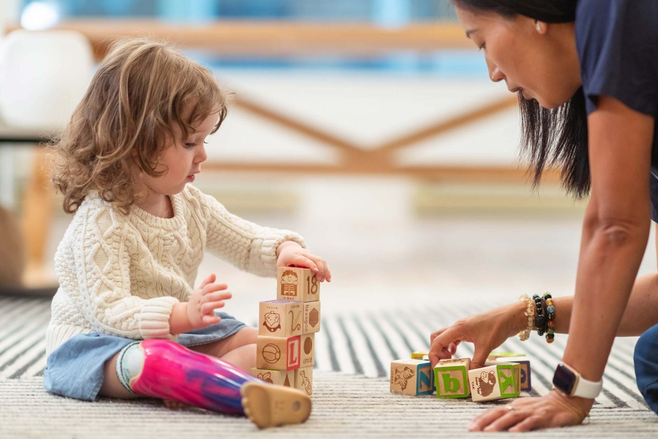curly haired toddler in a cream colored sweater with a purple and pink tie-died prosthetic leg stacks blocks printed with the alphabet on the carpet with therapist in navy scrubs