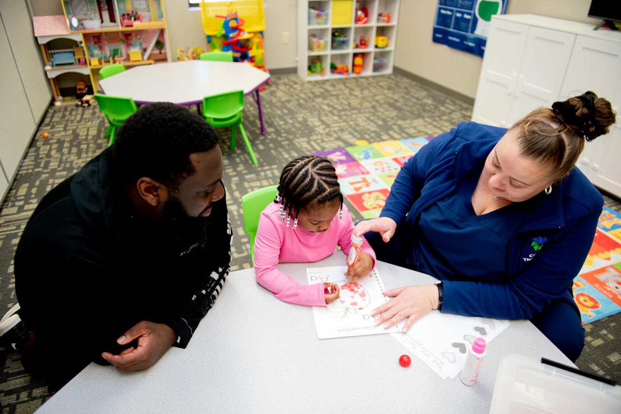 Young girl coloring at a table with her father and a preschool teacher at Carolina Therapeutics Academy in Rock Hill, SC